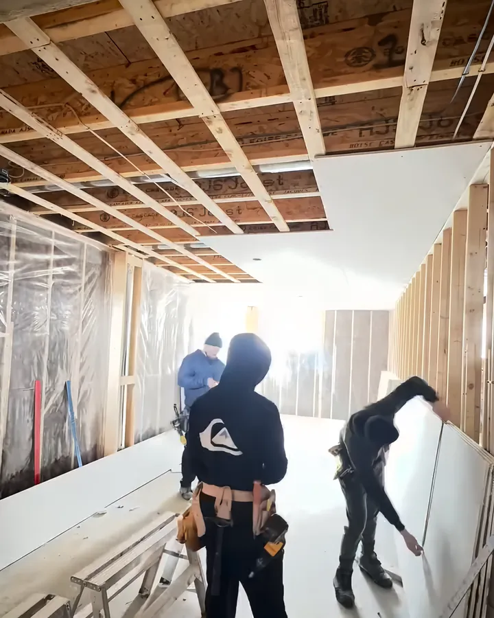 The KOSO crew hanging drywall in a new basement: one measuring a sheet, the ceiling half boarded.